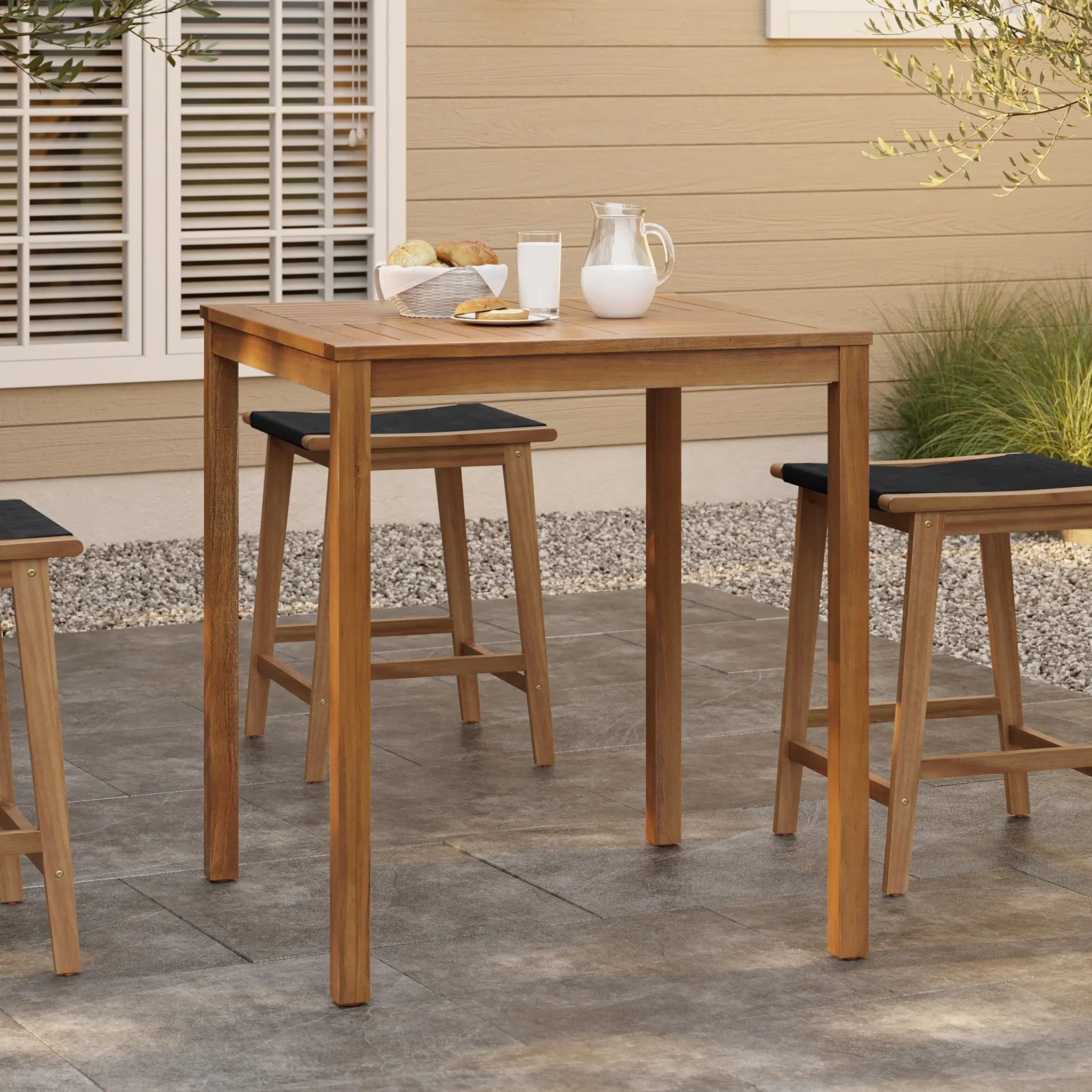 An outdoor acacia wood dining table, FSC-certified, holds a glass of milk, a jug, and a basket of bread. Surrounded by chairs on a stone patio with a beige building in the background, it offers a perfect setting for memorable meals and gatherings.