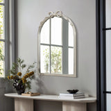 The decorative arched silhouette of the Ribbon Wall Mirror, complete with a charming bow detail, complements the light wood console table. A black vase with yellow flowers and a book adorns the table, while natural light pours in from a large nearby window.