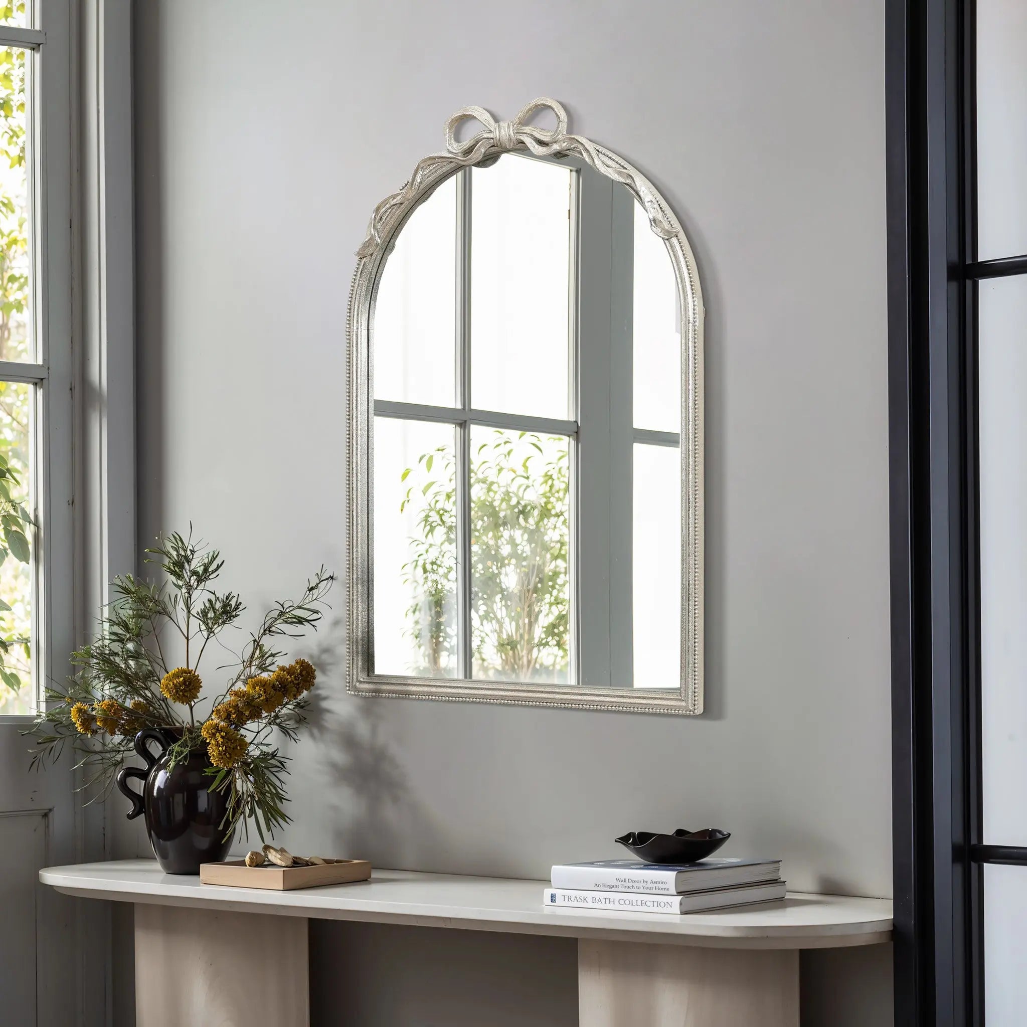 The decorative arched silhouette of the Ribbon Wall Mirror, complete with a charming bow detail, complements the light wood console table. A black vase with yellow flowers and a book adorns the table, while natural light pours in from a large nearby window.