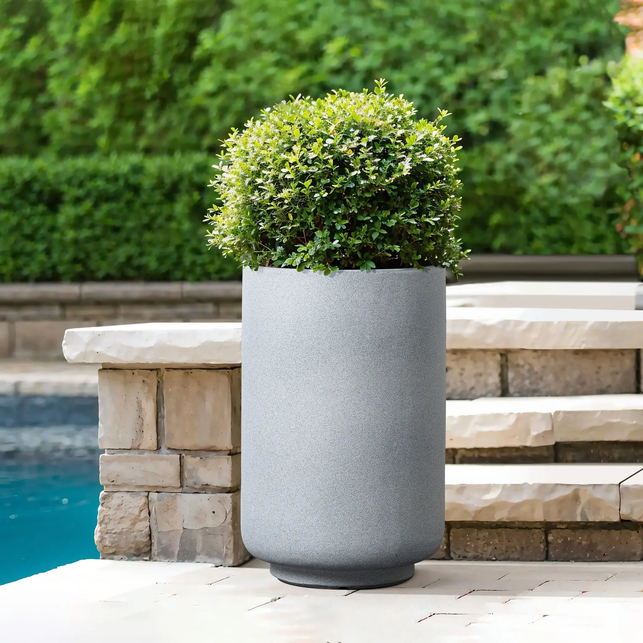 A modern minimalist decor touch, this gray round planter features a neatly trimmed bush, elegantly positioned on stone steps beside a shimmering pool. Surrounded by lush greenery, the weather-resistant planter adds contemporary charm to the serene setting.