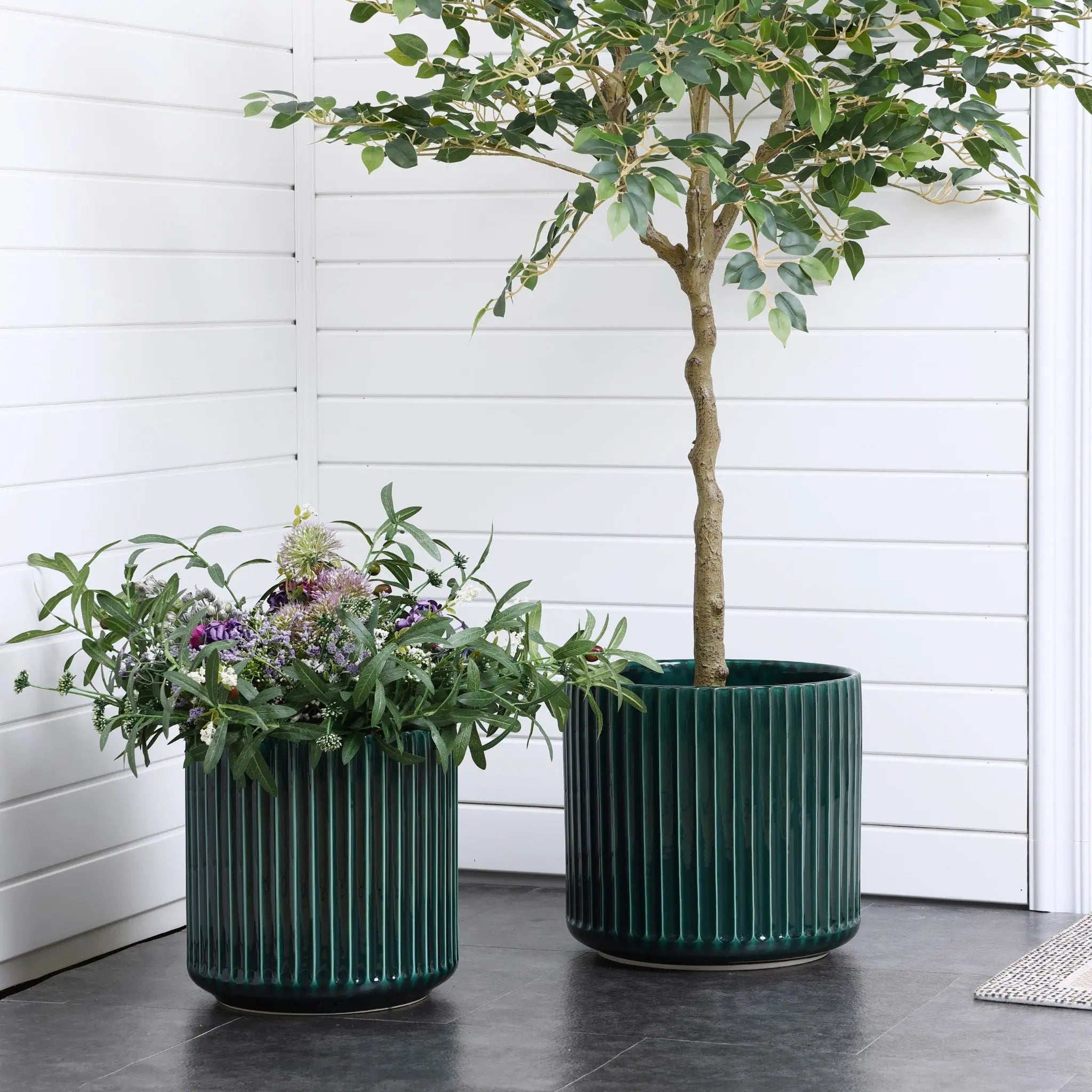 Two LuxenHome Jade green ribbed glazed round ceramic planters, 13 inches high, sit on a dark floor against white paneled walls. The left planter overflows with lush foliage and purple flowers, while the right holds a slender tree with translucent green leaves, crafting a modern plant paradise.