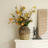 Rustic terracotta vase with yellow flowers and oranges on a mantel, next to a small sculpture of two figures walking, and a piece of textured wall art. The handcrafted weathered finish adds charm to this cozy setting.