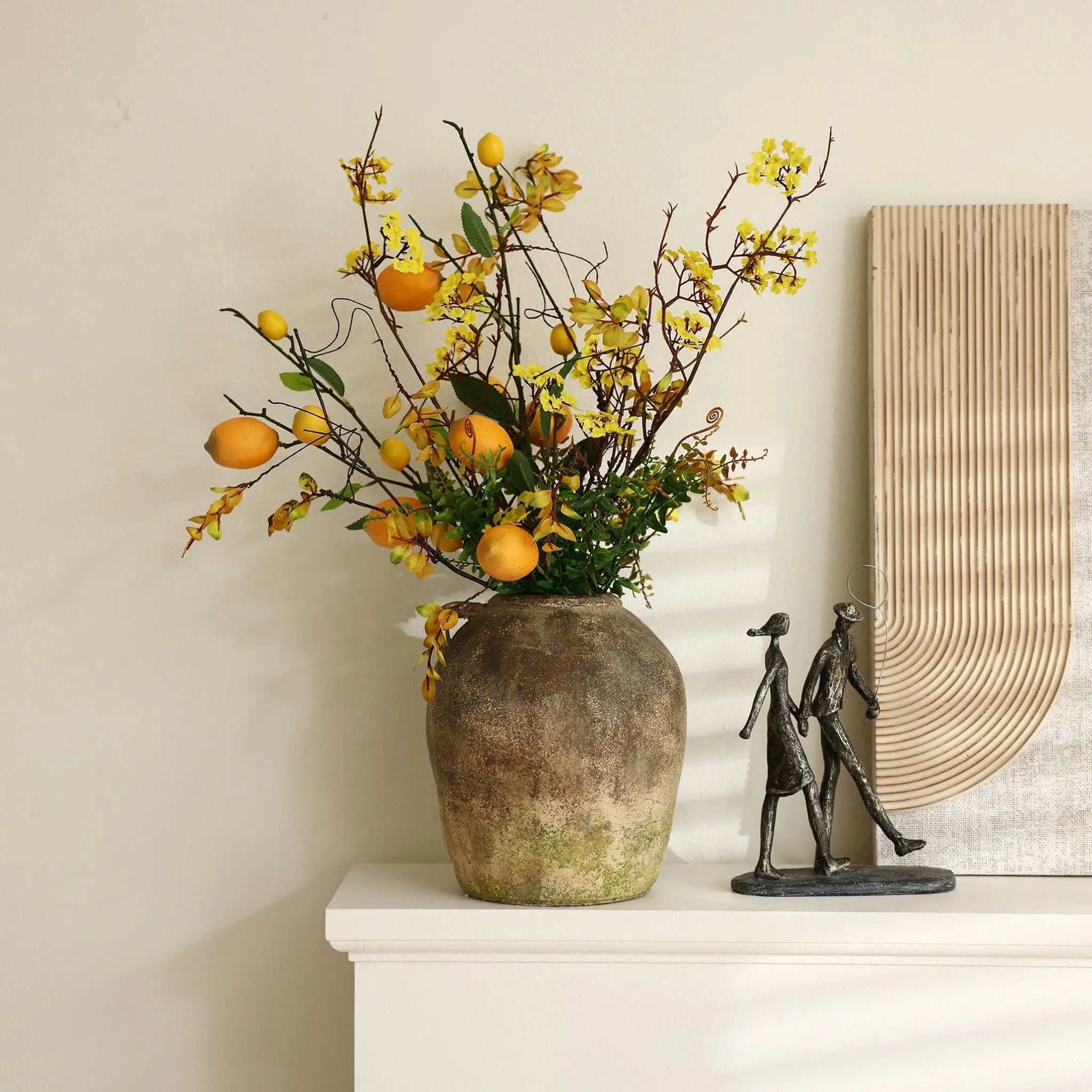 Rustic terracotta vase with yellow flowers and oranges on a mantel, next to a small sculpture of two figures walking, and a piece of textured wall art. The handcrafted weathered finish adds charm to this cozy setting.