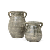 Two handcrafted, rustic green-gray clay pots with handles, one larger and the other smaller, stand side by side against a plain white background. These vessels evoke a sense of historic depth reminiscent of ancient terracotta jug vases.