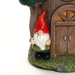 A garden gnome figurine with a red hat and white beard stands next to a charming gnome tree house with a wooden door, making it a delightful addition to your outdoor space decor.