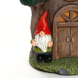 A garden gnome figurine with a red hat and white beard stands next to a charming gnome tree house with a wooden door, making it a delightful addition to your outdoor space decor.