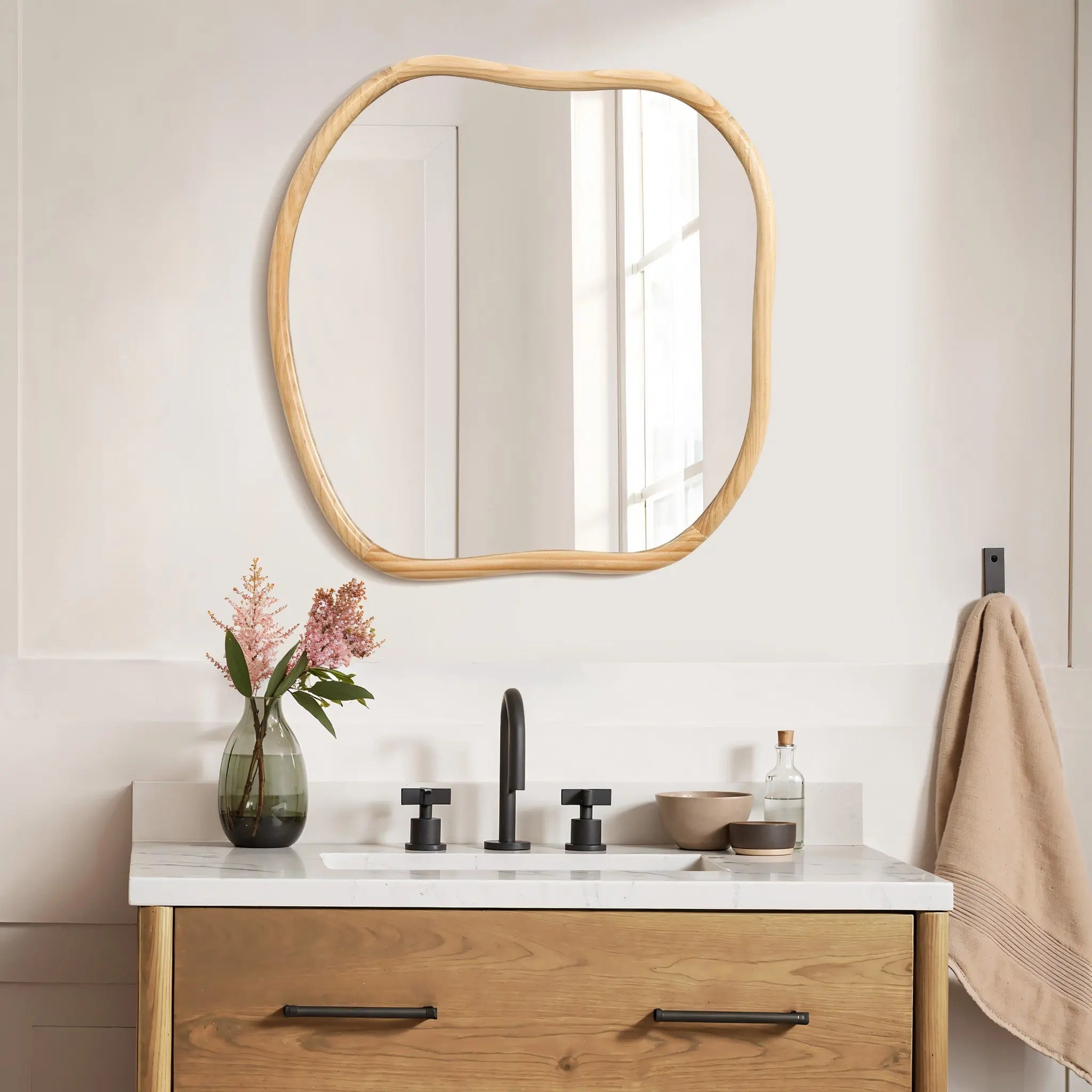 The bathroom features a stylish wooden vanity and an irregular-shaped wall mirror with a natural pine frame. A black faucet complements the decor, while a vase of flowers and a beige towel rest elegantly on the counter, bathed in natural light streaming through the window.
