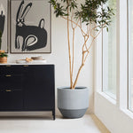 A tall modern minimalist planter stands beside a black cabinet with gold handles. Ceramic bowls and plates rest on top, while rabbit-themed artwork hangs above. Large windows flood the space with natural light, enhancing the indoor and outdoor decor appeal.