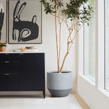 A tall modern minimalist planter stands beside a black cabinet with gold handles. Ceramic bowls and plates rest on top, while rabbit-themed artwork hangs above. Large windows flood the space with natural light, enhancing the indoor and outdoor decor appeal.