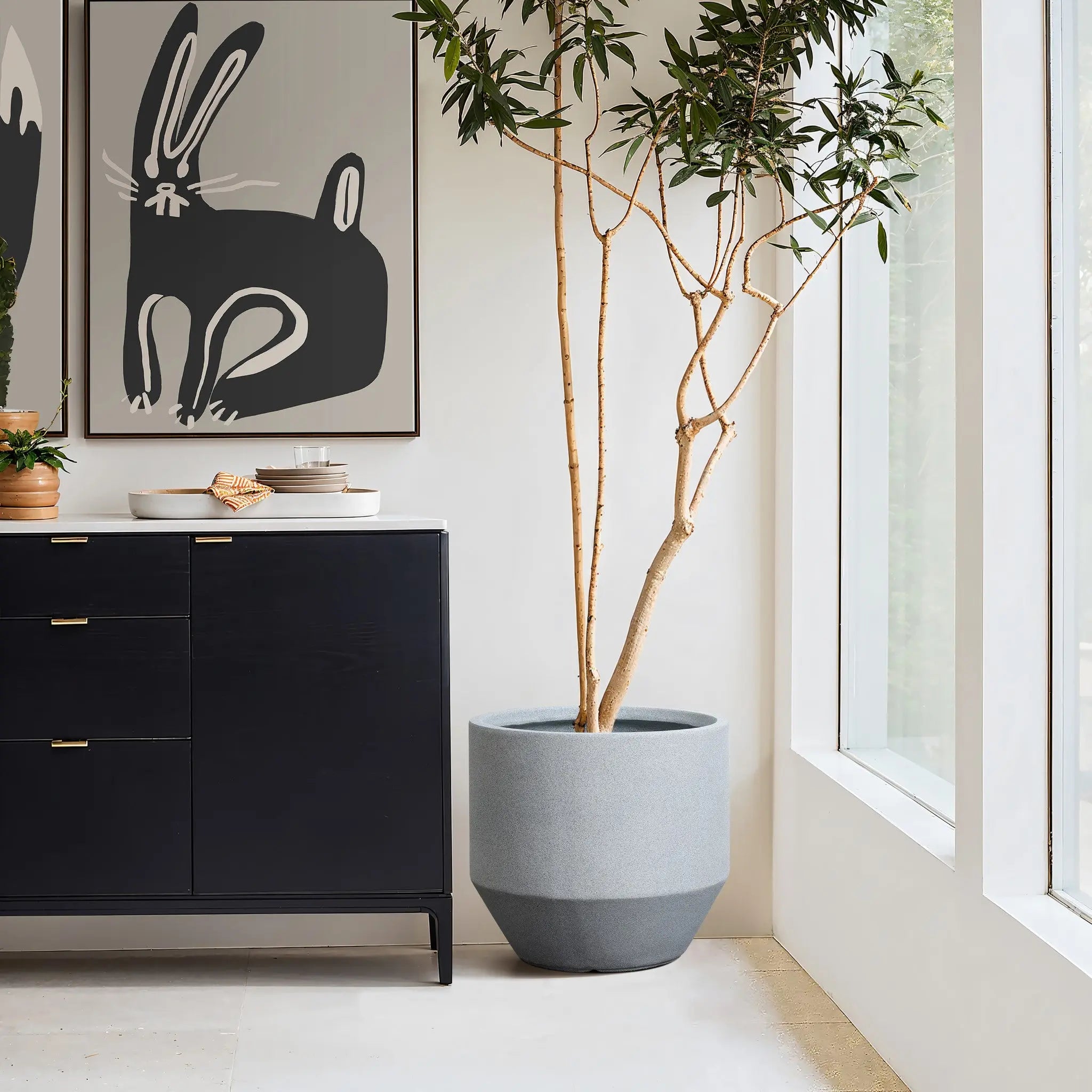 A tall modern minimalist planter stands beside a black cabinet with gold handles. Ceramic bowls and plates rest on top, while rabbit-themed artwork hangs above. Large windows flood the space with natural light, enhancing the indoor and outdoor decor appeal.