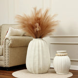 A large, modern white vase with brown pampas grass sits as an artistic centerpiece next to a smaller, textured white vase on a beige rug near a beige armchair.