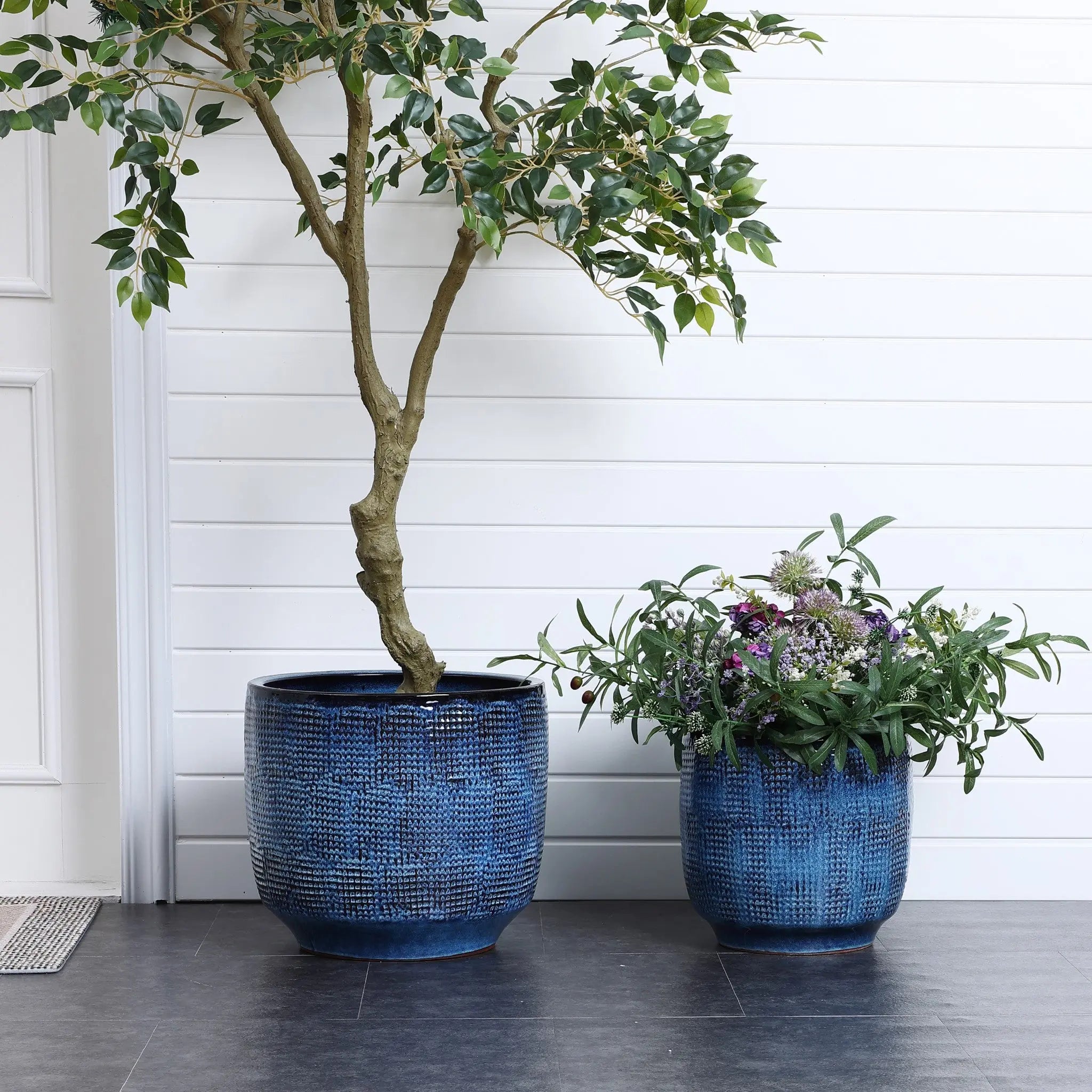 Two LuxenHome Ocean blue glazed round ceramic planters rest on a dark floor against a white paneled wall. The left one contains a tall tree with green leaves, while the right one showcases flowering plants with purple and white blooms.
