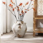 A handcrafted vintage vase with orange flowers and branches graces a wooden floor, complementing the rustic charm of a wooden cabinet.