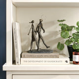 A modern resin sculpture of two abstract figures with a balloon elegantly stands on a stack of books, nestled next to a potted plant and a candle on the shelf.