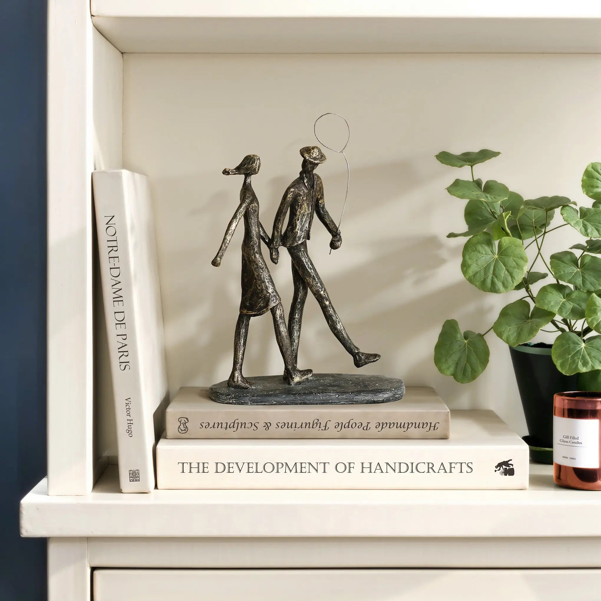A modern resin sculpture of two abstract figures with a balloon elegantly stands on a stack of books, nestled next to a potted plant and a candle on the shelf.