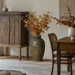 A stoneware vase with autumnal foliage sits on a wooden floor next to a wicker chair and wooden table, exuding rustic charm, with a cabinet and small jug in the background.