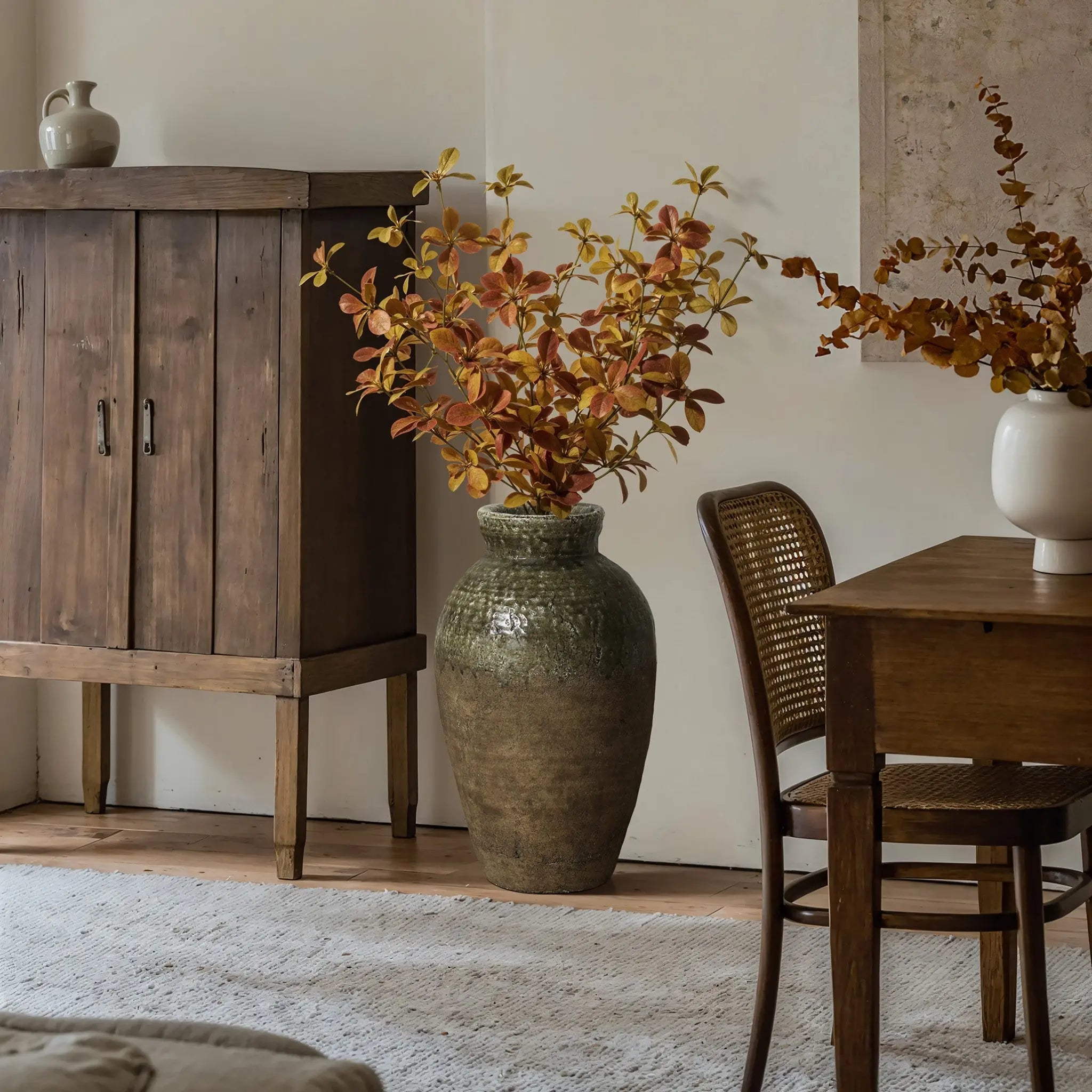 A stoneware vase with autumnal foliage sits on a wooden floor next to a wicker chair and wooden table, exuding rustic charm, with a cabinet and small jug in the background.