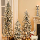 Three pre-lit artificial trees with lifelike PVC branches, snow-frosted and glowing, stand beside a fireplace and window in a living room, with wrapped gifts and a decorative reindeer creating a winter wonderland scene.