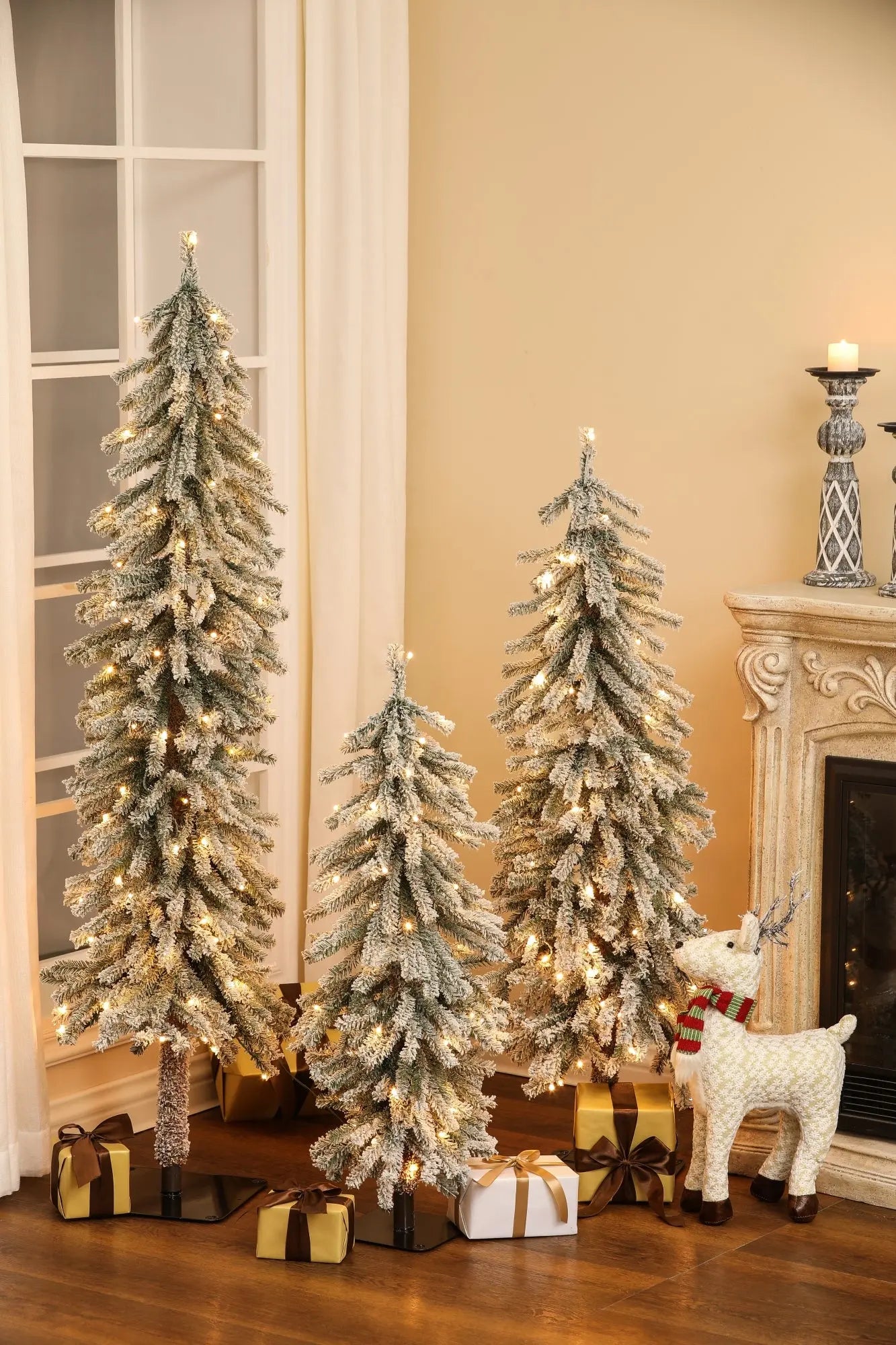 Three pre-lit artificial trees with lifelike PVC branches, snow-frosted and glowing, stand beside a fireplace and window in a living room, with wrapped gifts and a decorative reindeer creating a winter wonderland scene.
