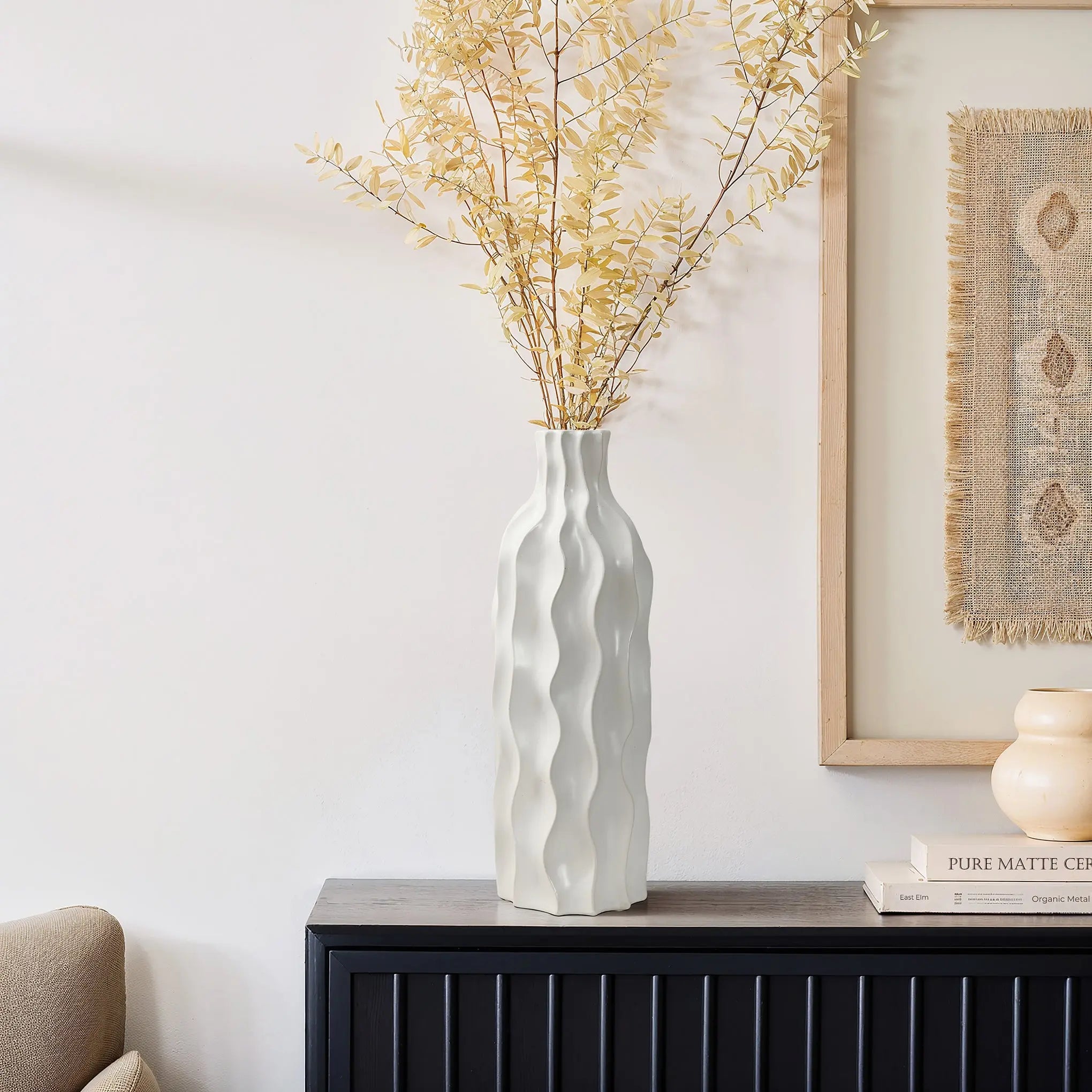 A tall, wavy ceramic vase with an elegant design holds dried branches on a black console table, next to a framed artwork and two books. This versatile piece adds sophistication to any room.
