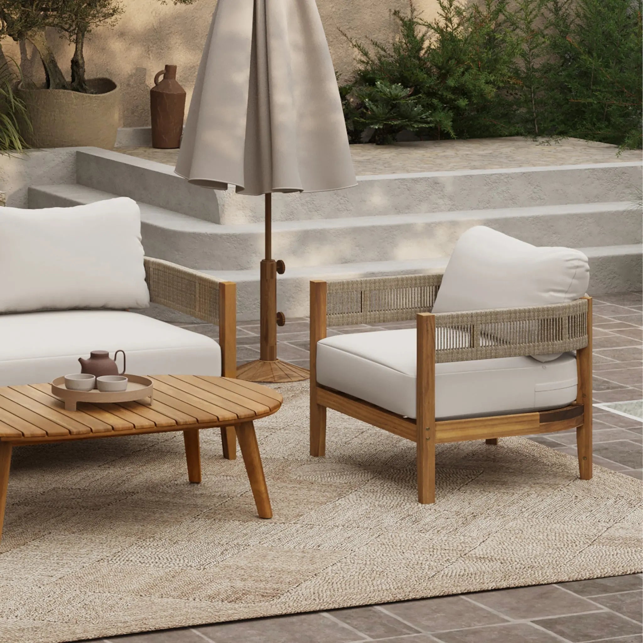 Outdoor patio with Club-Style Patio Furniture, featuring a wooden coffee table, cushioned chairs, a neutral rug, and a closed umbrella on tiled flooring with lush plants in the background.