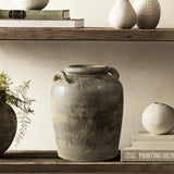 The LuxenHome Fou antique terracotta vase, a rustic handcrafted piece with handles, is showcased on a wooden shelf surrounded by small white textured ornaments and greenery. Stacked books with muted covers below the shelf enhance the minimalist decor.