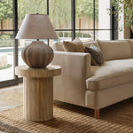 A modern living room features a beige sofa with decorative pillows. A LuxenHome dark oak finish fluted round side table holds a textured lamp. Large windows show an outdoor view, while a woven rug covers the light wood floor, and pillows add accents to the neutral decor.