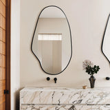 An irregular oval mirror with a black frame hangs above a marble sink with sleek black fixtures, adding to the modern aesthetic. A vase with flowers and a small dish sit gracefully on the counter.