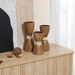Three wooden hourglass-shaped candle holders with a rattan design and twine sit gracefully on a wooden table alongside decor objects. A textured art piece serves as the backdrop.