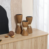 Three wooden hourglass-shaped candle holders with a rattan design and twine sit gracefully on a wooden table alongside decor objects. A textured art piece serves as the backdrop.