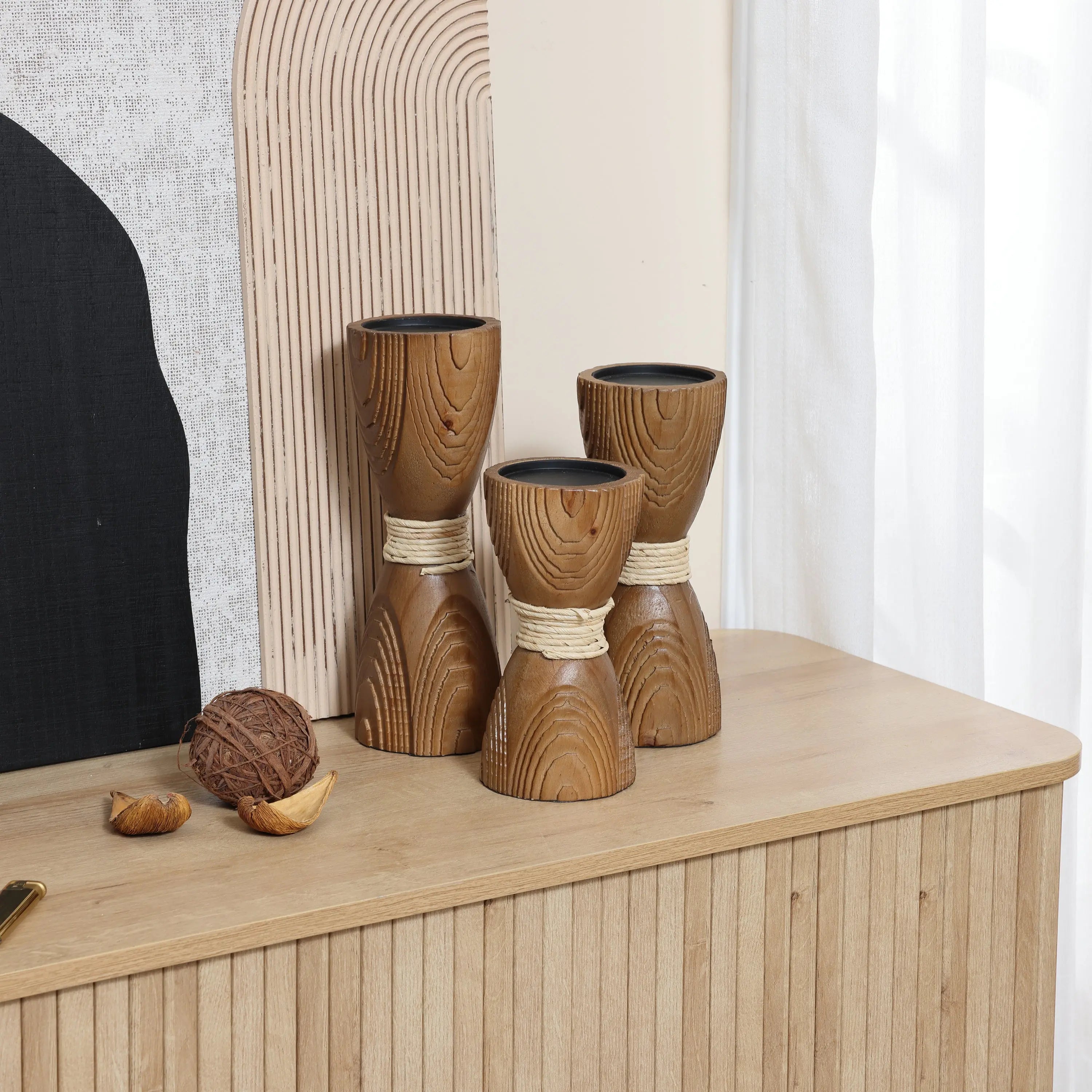 Three wooden hourglass-shaped candle holders with a rattan design and twine sit gracefully on a wooden table alongside decor objects. A textured art piece serves as the backdrop.