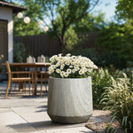 A large gray planter, featuring a marble ribbed texture and blooming white daisies, rests on the patio. In the background, an outdoor table and chairs are nestled among lush greenery and trees.