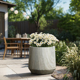 A large gray planter, featuring a marble ribbed texture and blooming white daisies, rests on the patio. In the background, an outdoor table and chairs are nestled among lush greenery and trees.