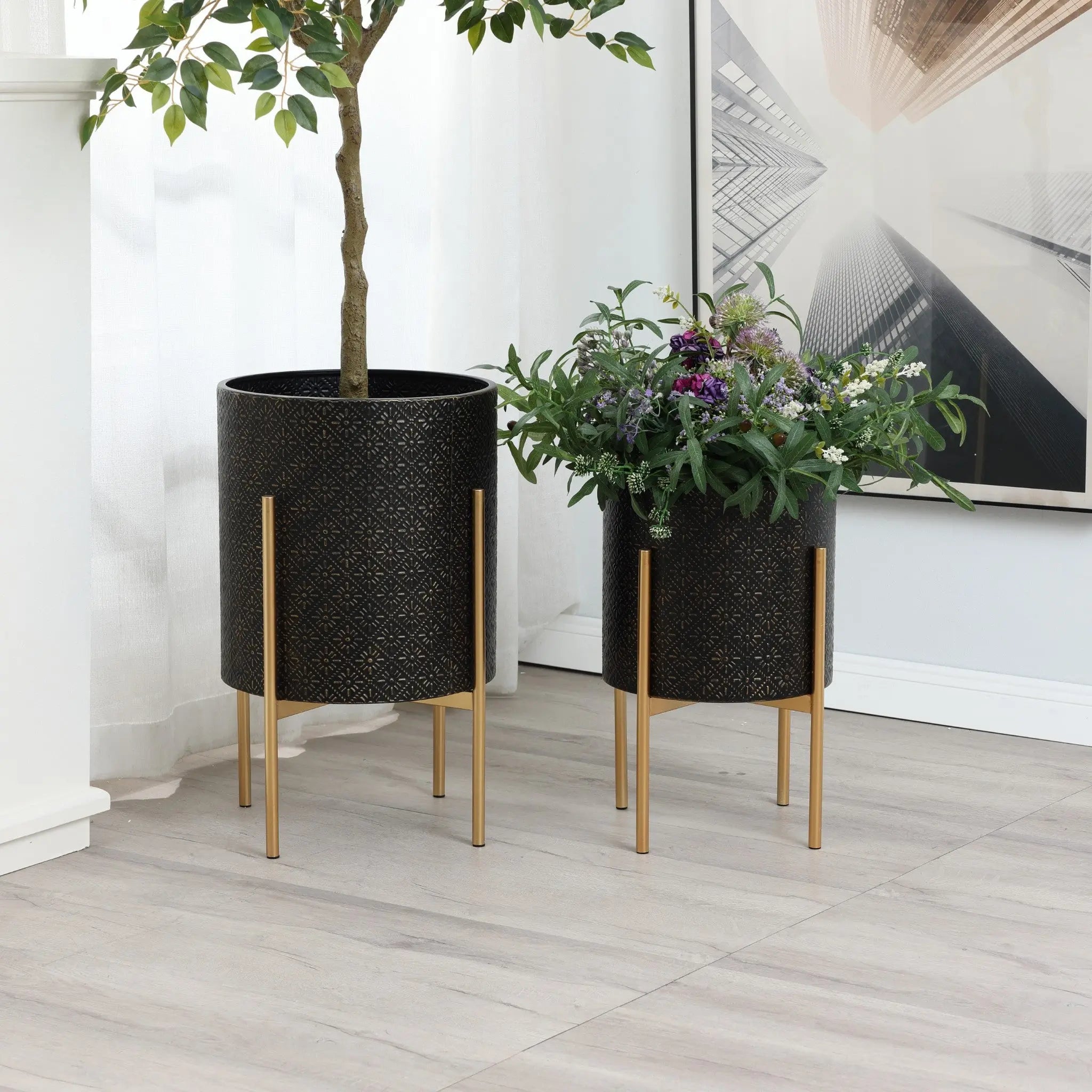 Two black plant pots with an elegant gold metal stand cradle a small tree and assorted flowers, resting on a light wood floor near a white wall adorned with modern framed artwork.