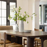 A dining table with a rustic charm features a white vase holding white flowers, a matching milk jug, and stacked plates. Four woven chairs surround the table, set in a bright room with large windows.