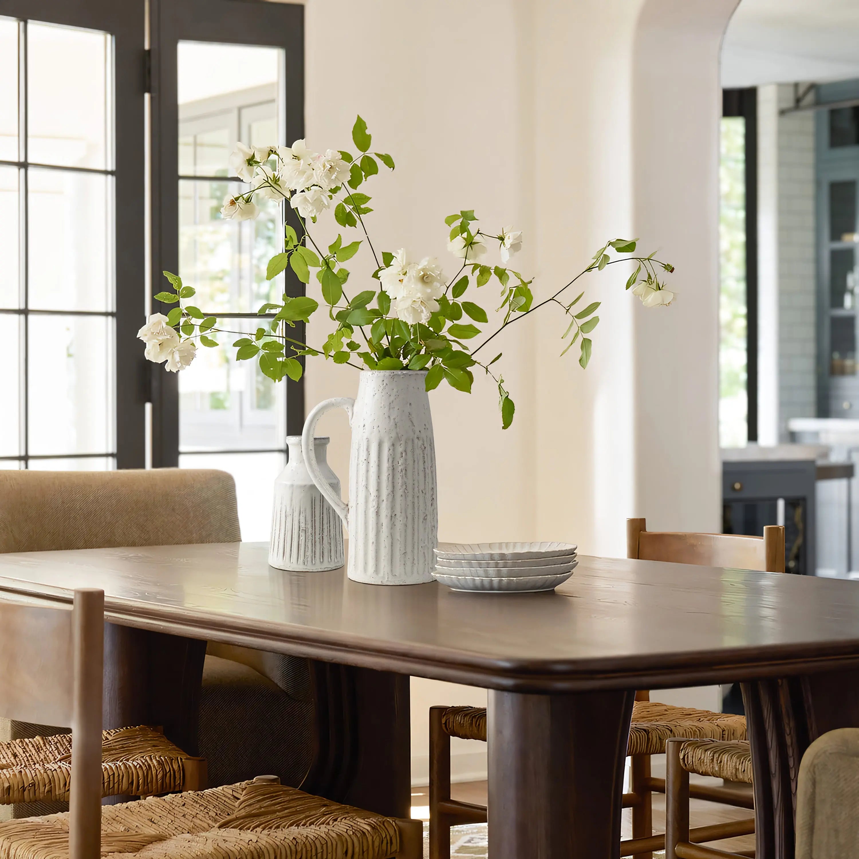 A dining table with a rustic charm features a white vase holding white flowers, a matching milk jug, and stacked plates. Four woven chairs surround the table, set in a bright room with large windows.