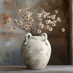 A LuxenHome rustic gray and white terracotta vase with handles, standing 11 inches high, holds a bouquet of dried white flowers on a wooden surface. The weathered background enhances the contemporary elegance of the vintage scene.