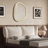 A bedroom featuring a beige upholstered bed with white and gray bedding exudes elegance. An oval wall mirror with a free-form frame hangs above the bed, complementing the abstract wall art and adding a touch of modern sophistication to the home decor.
