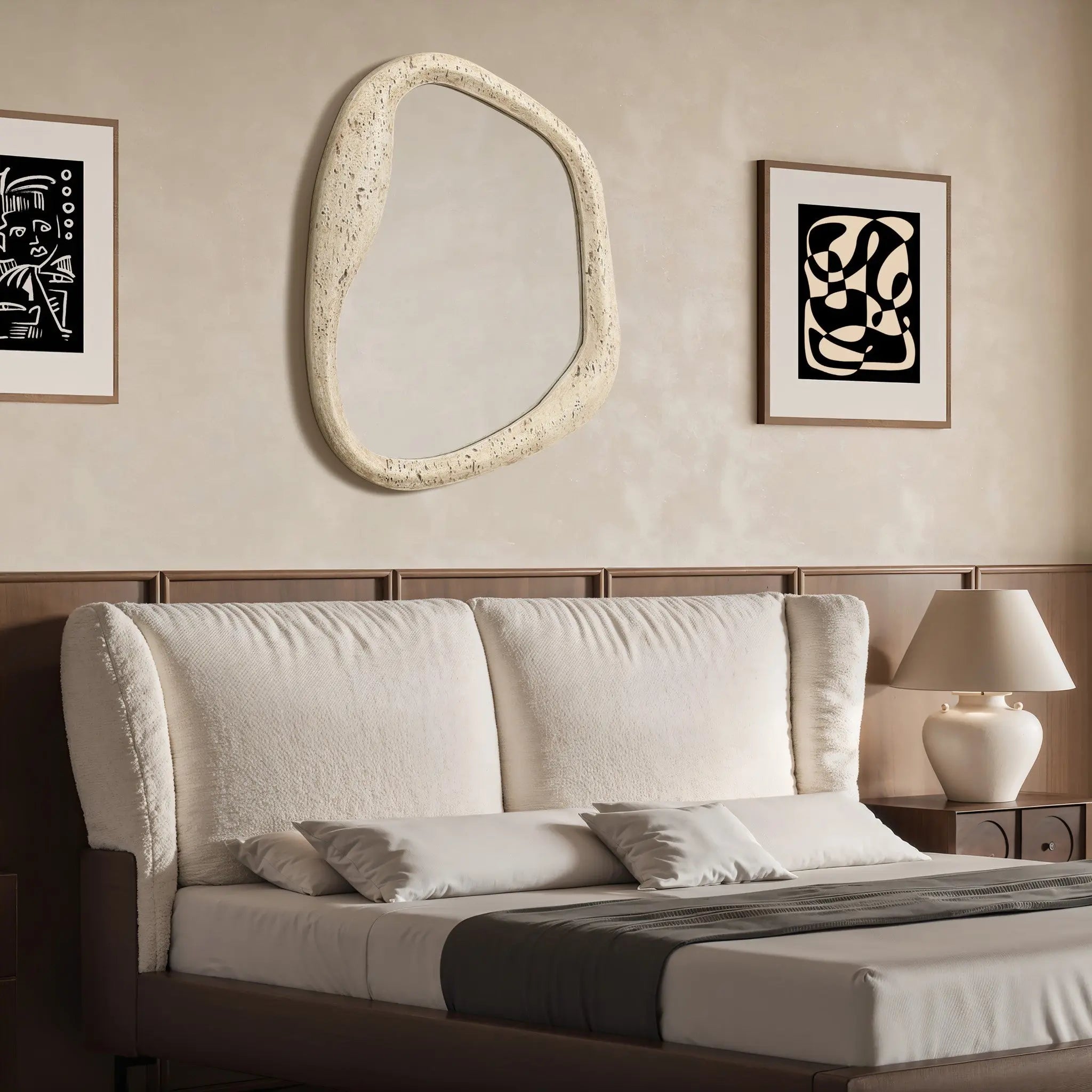 A bedroom featuring a beige upholstered bed with white and gray bedding exudes elegance. An oval wall mirror with a free-form frame hangs above the bed, complementing the abstract wall art and adding a touch of modern sophistication to the home decor.