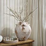 A large, handcrafted vintage vase with branches sits on a wooden table beside a tray with a carafe and two glasses, against a paneled wall. This versatile home decor piece adds rustic charm to any space.