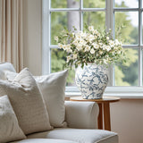 A handcrafted floral ceramic vase with white flowers and greenery sits on a wooden table next to a white sofa.