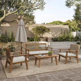 Outdoor furniture set featuring two chairs, a loveseat, and a table on a rug. A closed umbrella stands nearby, enhancing the minimalist design. The scene is complemented by plants, trees, and a stone wall under a clear sky. Made from FSC-certified Acacia wood for eco-friendly elegance.