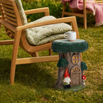 A small multifunctional table shaped like a tree trunk with a gnome and mushroom design stands beside a cushioned wooden chair on grass, with a white mug on top¡ªperfect for adding charm to your outdoor space decor.