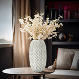 A LuxenHome Modern geometric pattern resin tall vase, 13.5 h, with an antique effect in white displays a bouquet of white cherry blossoms. It rests on a round wooden table in a cozy room with draped curtains, a brown accent chair, and shelves in the background.