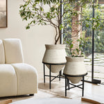 Two large cachepot planters in beige containers on black stands, elegantly placed near a modern white sofa by a large window with a garden view, adding a touch of indoor space decor charm.