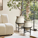 Two large cachepot planters in beige containers on black stands, elegantly placed near a modern white sofa by a large window with a garden view, adding a touch of indoor space decor charm.