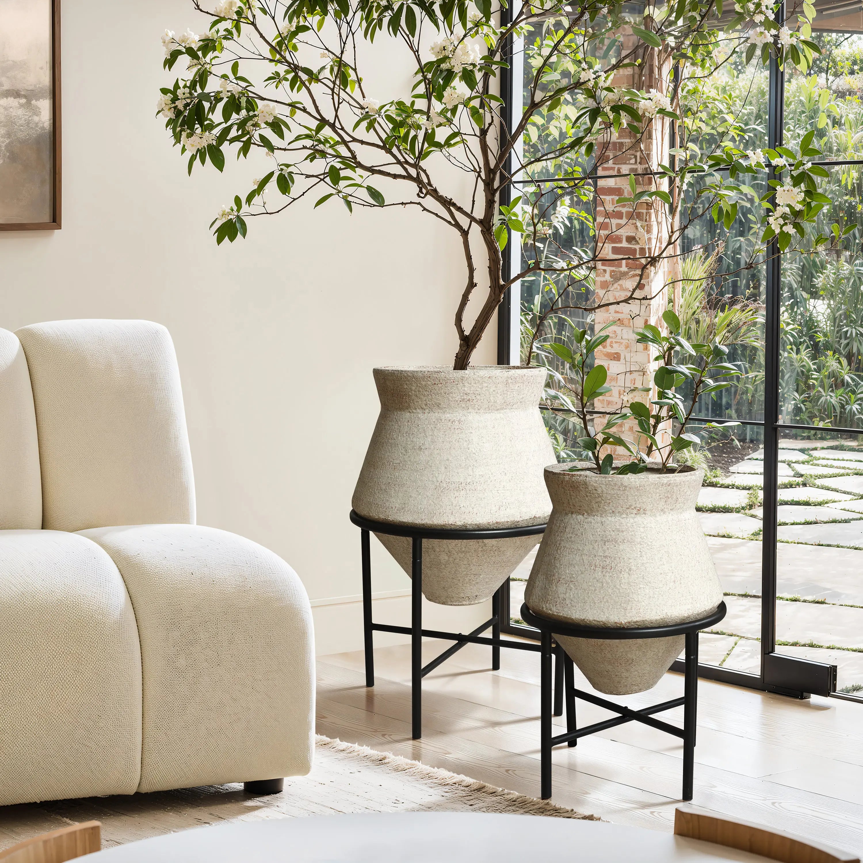 Two large cachepot planters in beige containers on black stands, elegantly placed near a modern white sofa by a large window with a garden view, adding a touch of indoor space decor charm.