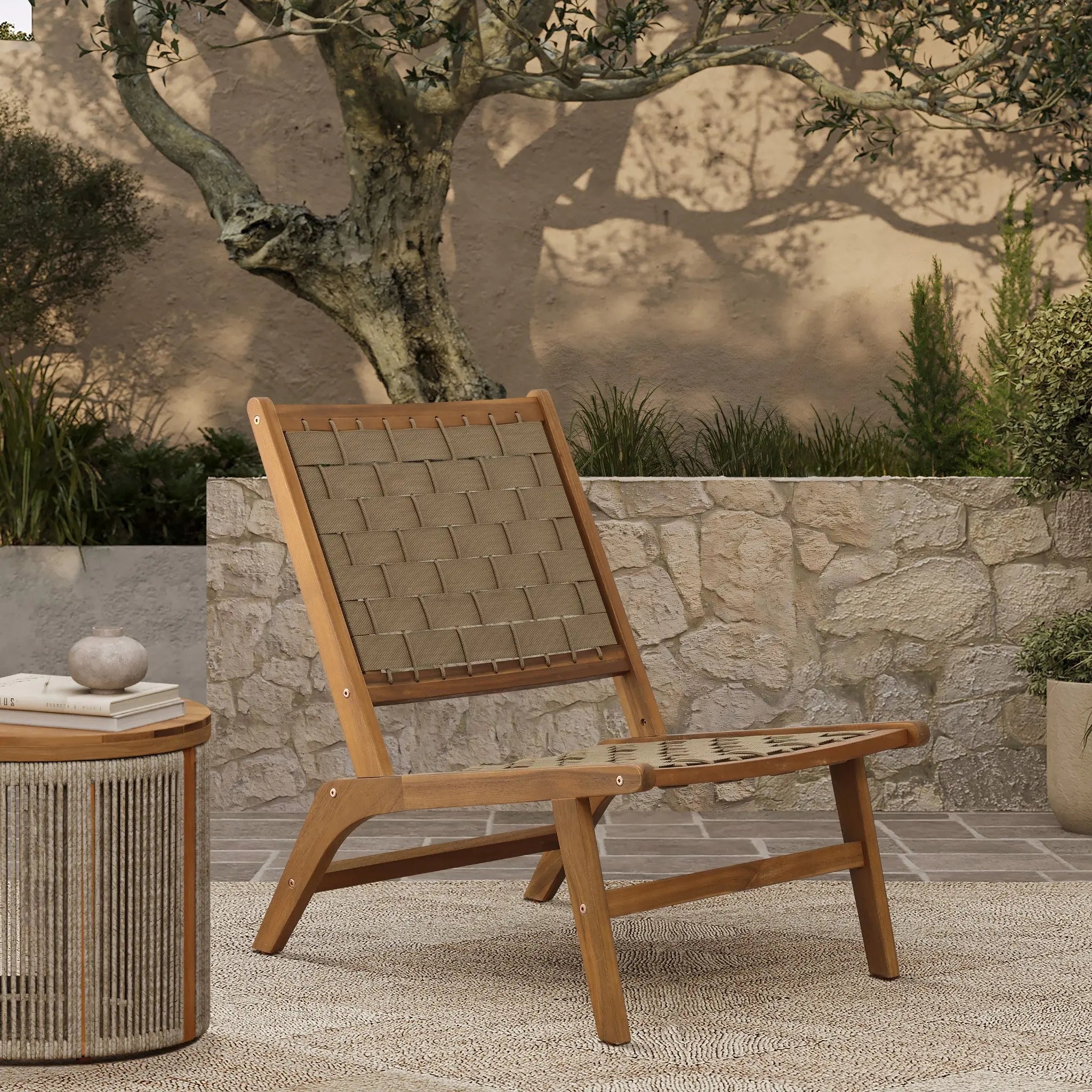 An Acacia wood chair with a woven seat and backrest sits on a textured rug beside a round table. In the background, the stone wall and tree cast intricate shadows, enhancing this outdoor furniture settings natural charm.