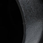 Close-up of a textured, curved, dark green metal surface featuring a unique white irregular pattern, with a small circular detail, set against a black background.