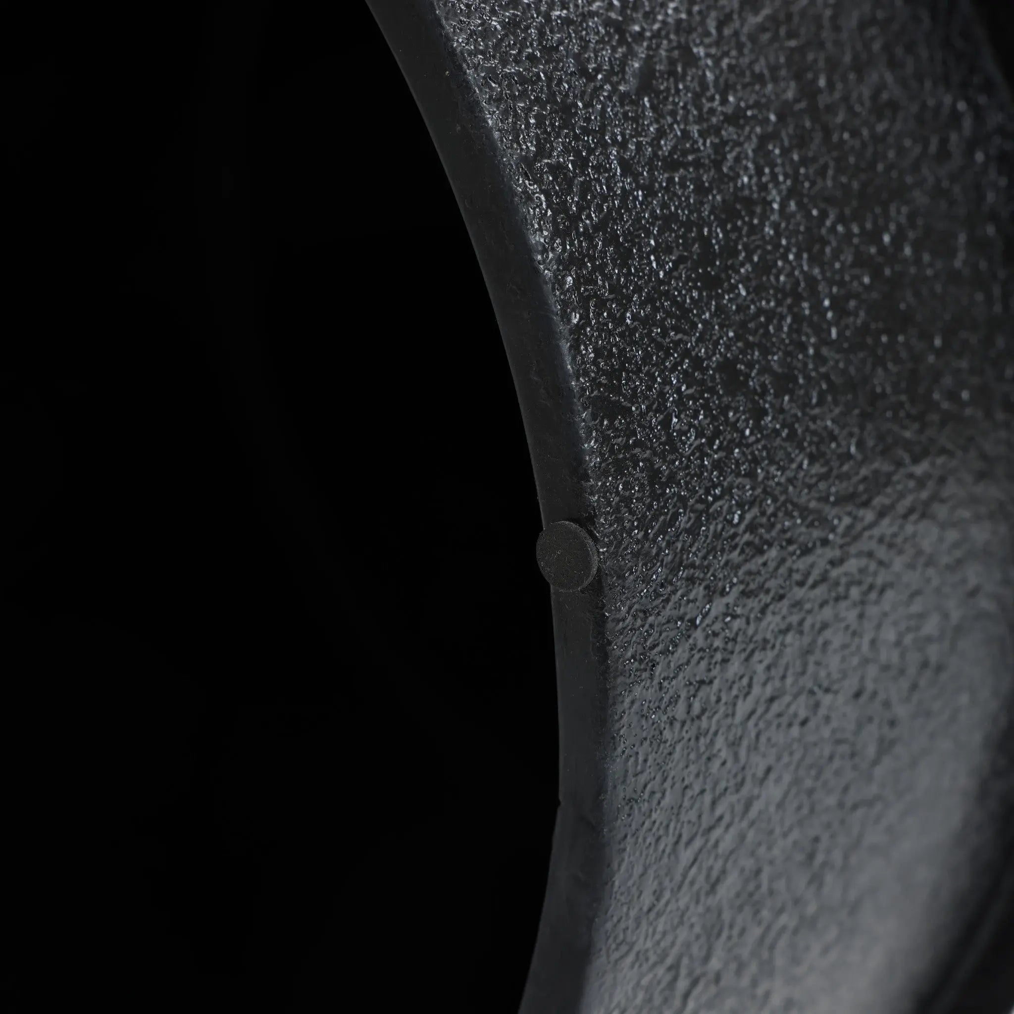 Close-up of a textured, curved, dark green metal surface featuring a unique white irregular pattern, with a small circular detail, set against a black background.